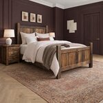 Diagonal angled room view showing the right side of a farmhouse panel bed with a plank footboard, in a plum-paneled bedroom.
